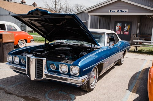 A classic blue convertible car with the hood open, parked in front of a fair office building.