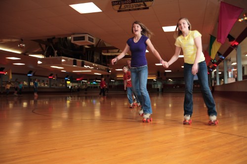 Two girls roller skating hand in hand in a lively indoor rink, with others skating in the background.