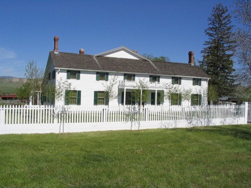 A large white house with green shutters, surrounded by a white picket fence and green grass under a clear blue sky.