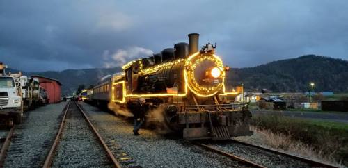 A decorated steam train with lights, parked on tracks with mountains in the background under a cloudy sky.