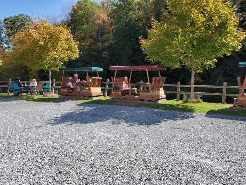 A sunny outdoor area with wooden swings and trees, surrounded by gravel and a wooden fence.