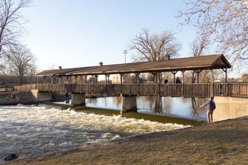 A wooden bridge spans a river, with people walking on it and along the bank, surrounded by trees and a clear sky.