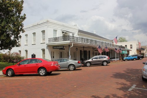 Historic building with a balcony, surrounded by parked cars and American flags, on a brick street under a cloudy sky.