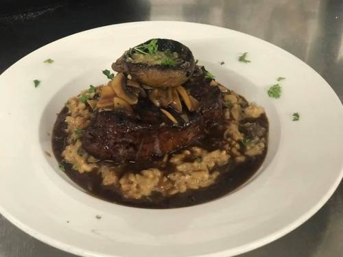A plated dish featuring a grilled steak on a bed of risotto, topped with mushrooms and garnished with herbs.
