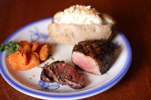 A plate with grilled steak, sliced meat, carrots, and a baked potato topped with sour cream.