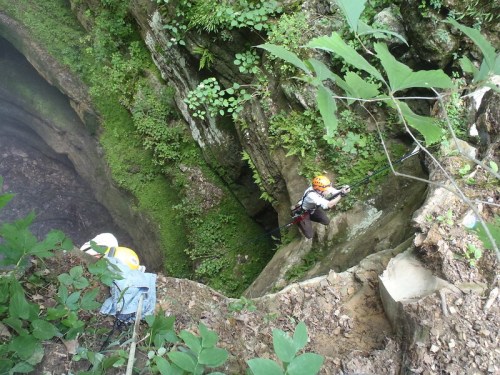 Two climbers navigate a rocky canyon surrounded by lush greenery, one climbing down while the other rests nearby.