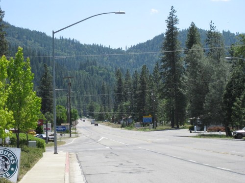 A quiet road lined with trees, leading into a mountainous area under a clear blue sky.