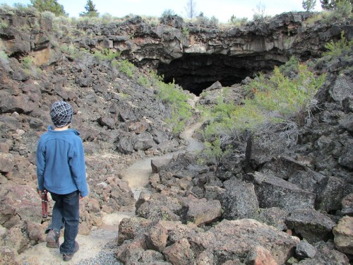 A child stands on a rocky path leading to a dark cave entrance surrounded by sparse vegetation.