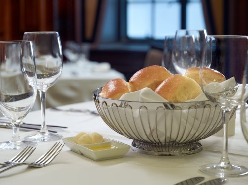A silver bowl of freshly baked rolls sits on a elegantly set dining table with glassware and utensils.