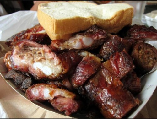 A pile of smoked meats, including ribs and sausage, topped with a slice of bread on a paper-lined tray.