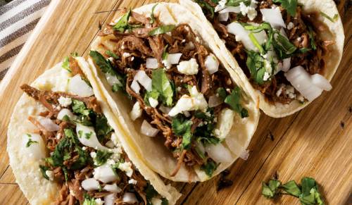 Three tacos filled with shredded meat, onions, cilantro, and cheese, served on a wooden cutting board.