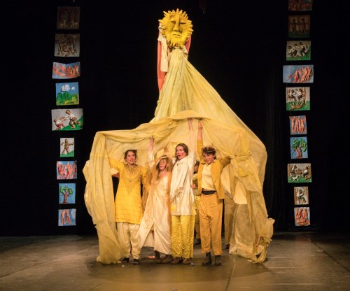 Four performers in yellow costumes hold up a large, draped figure resembling a lion, set against a colorful backdrop.