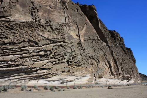 A rugged rock formation with layered textures under a clear blue sky, bordered by a fence and sparse vegetation.