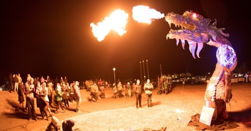 A large dragon sculpture breathes fire as a crowd watches at night, illuminated by colorful lights.
