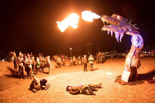A dragon sculpture breathes fire as a crowd watches, with performers on the ground in a nighttime outdoor setting.
