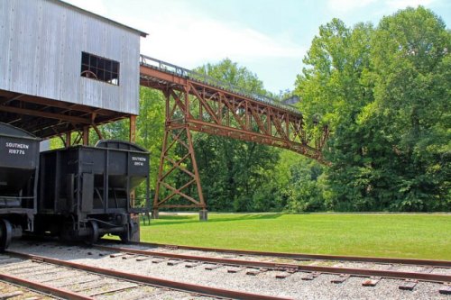 A rusted train trestle spans a green landscape, with black train cars parked on the tracks below.