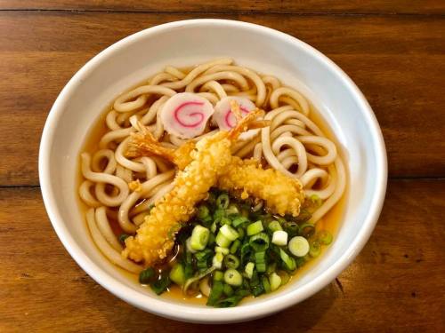 A bowl of udon noodles topped with tempura shrimp, green onions, and pink fish cakes in a savory broth.