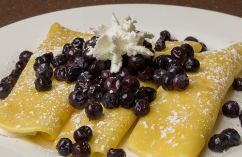 Three crepes topped with fresh blueberries and whipped cream, dusted with powdered sugar on a white plate.