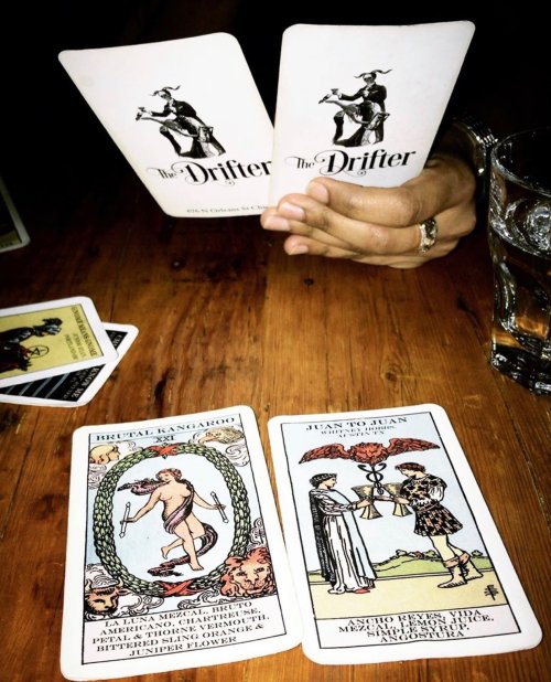 A person holds two playing cards labeled "The Drifter," with two additional cards on a wooden table and a glass nearby.