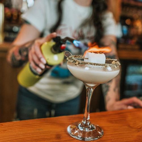 A bartender uses a torch to caramelize the top of a cocktail in a glass with a graham cracker rim.