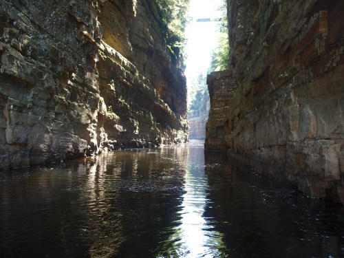 A serene canyon with steep rock walls and calm water reflecting sunlight. Lush greenery lines the top of the cliffs.