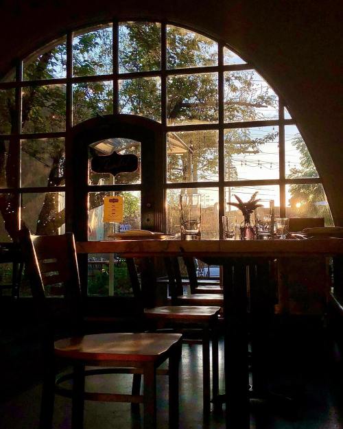 A cozy restaurant interior with empty chairs and a large window showcasing a sunset and trees outside.