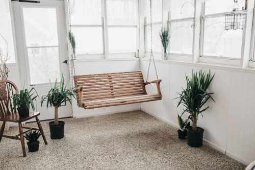 A wooden swing hangs in a bright room with potted plants and a chair, creating a cozy, inviting atmosphere.
