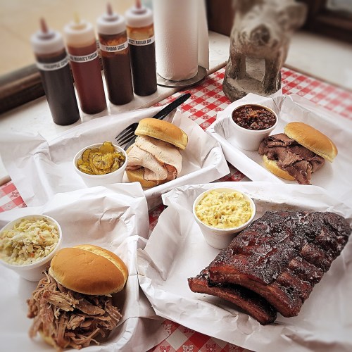 A spread of barbecue dishes including sandwiches, ribs, coleslaw, and sides, with sauces in the background.