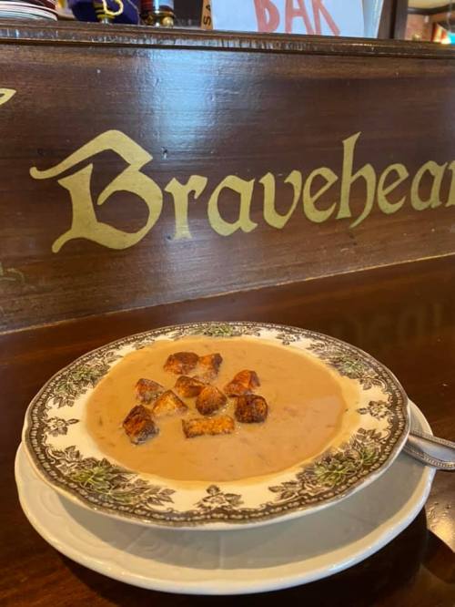 A bowl of creamy soup topped with croutons, placed on a decorative plate in front of a wooden bar with "Braveheart" sign.