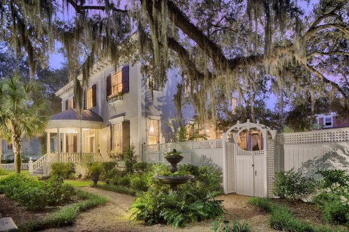 A charming two-story house with a porch, surrounded by lush greenery and a decorative gate, illuminated at dusk.