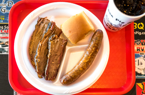 A plate with sliced brisket, a sausage, a bun, and a cup of soda on a red tray.