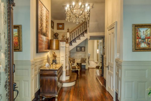 Elegant hallway with a chandelier, wooden floors, and decorative artwork, leading to a staircase and living area.