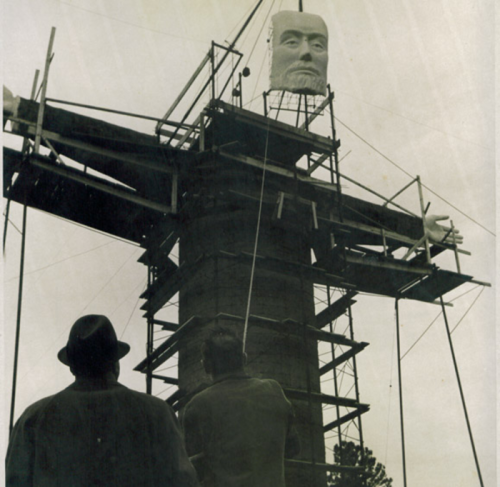 Workers observe the construction of a large statue on a tall structure, with scaffolding surrounding it.