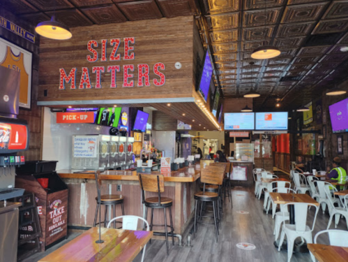 Interior of a casual restaurant with wooden decor, neon sign reading "SIZE MATTERS," and seating area.