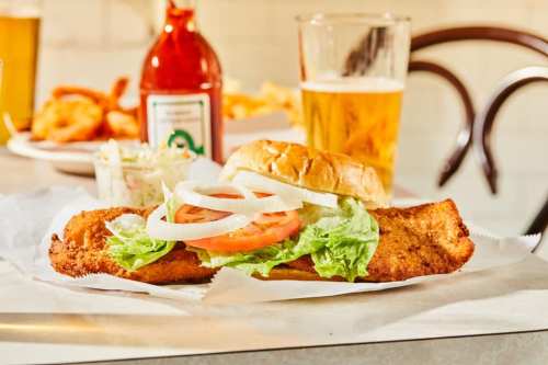 A crispy fish sandwich with lettuce, tomato, and onion, served with a side of coleslaw and a glass of beer.