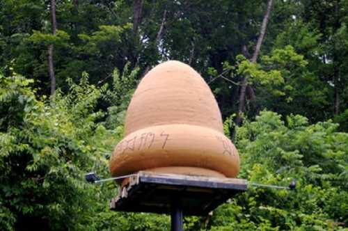 A large, clay sculpture resembling an egg or dome, placed on a wooden platform amidst lush green trees.