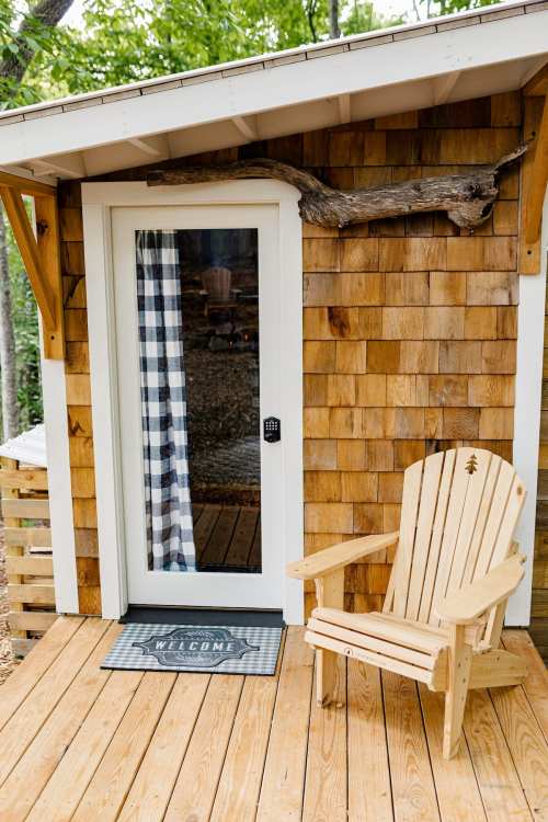 A cozy entrance with a wooden door, checkered curtain, and a welcoming mat beside a wooden chair on a deck.