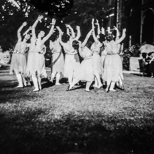 A group of dancers in flowing dresses performing in a garden, with arms raised and a vintage aesthetic.
