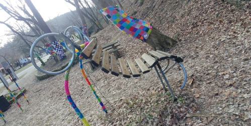 Colorful, knitted patterns adorn a unique, curved wooden bench in a park surrounded by bare trees and fallen leaves.