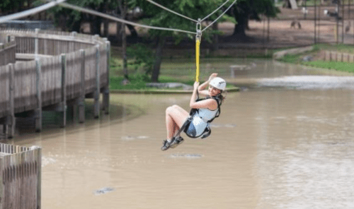 A person in a helmet zip-lining over a muddy river, surrounded by trees and wooden structures.
