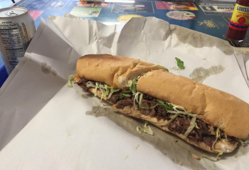 A partially eaten sub sandwich filled with beef and lettuce, resting on crumpled white paper, with a soda can nearby.