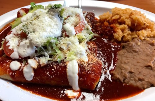 A plate of enchiladas topped with lettuce, tomatoes, cheese, and sour cream, served with rice and refried beans.