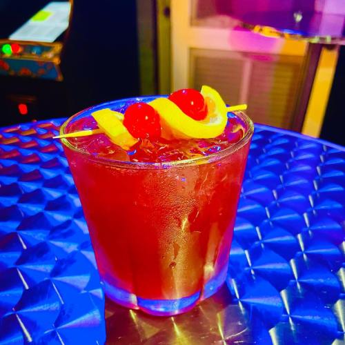 A colorful cocktail in a glass, garnished with cherries and a lemon twist, on a shiny blue surface.