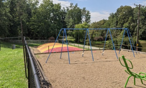 A playground with swings and a colorful slide, surrounded by green grass and trees under a clear blue sky.