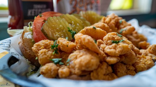 A close-up of a plate featuring crispy fried shrimp, lettuce, tomato, and pickles on a bun, garnished with parsley.