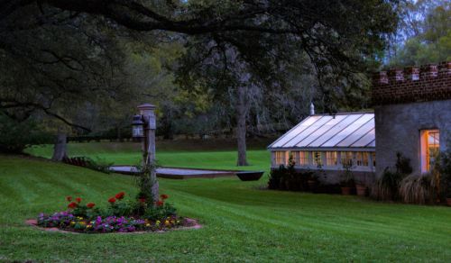 A serene garden scene at dusk, featuring a greenhouse, lush grass, and colorful flower beds surrounded by trees.