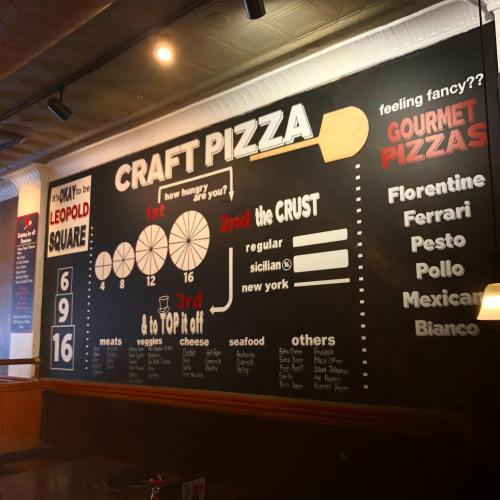 A menu board displaying options for crafting pizzas, including crust types and toppings, in a restaurant setting.