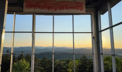View from a lookout tower, showcasing mountains and trees under a sunset sky with a drawing hanging on the wall.