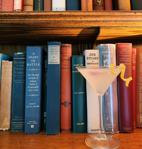 A cocktail sits on a table in front of a bookshelf filled with historical books.