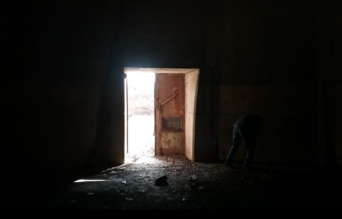 A dimly lit room with a partially open door, revealing bright light outside and a figure bending down near the floor.
